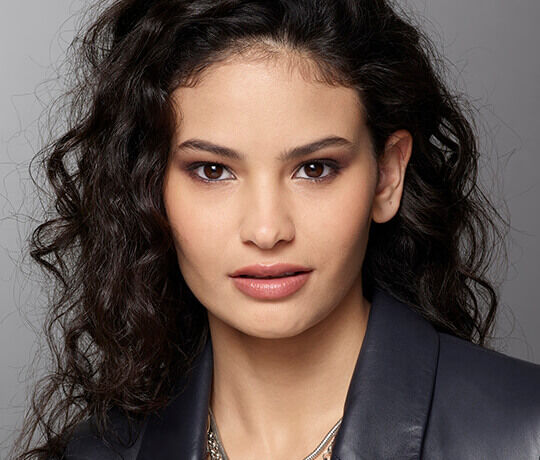 Close-up portrait of a young woman with curly hair, wearing a dark jacket, and displaying a subtle smile and natural makeup.