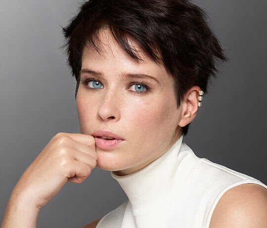 A young woman with short dark hair, wearing a high-neck top, poses thoughtfully with her chin resting on her hand, showcasing her striking blue eyes and subtle makeup against a gray background.