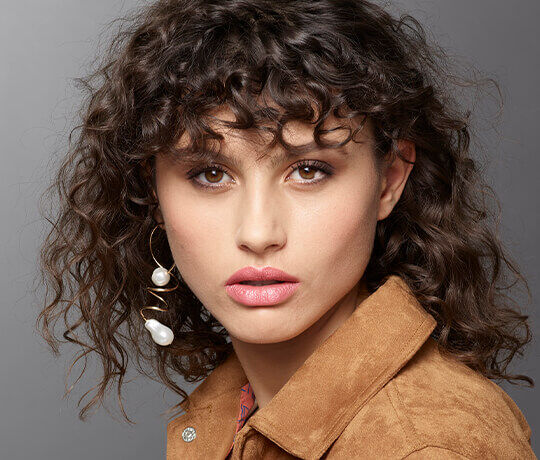 A young woman with curly hair wears a brown suede jacket and large, elegant earrings featuring pearls. She has a natural makeup look, showcasing her clear skin and defined features, while gazing directly at the camera.