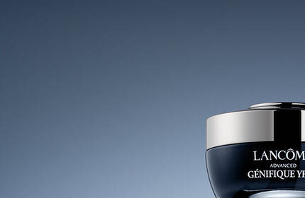 A dark gradient background features two Lancome skincare products: a jar labeled "Advanced Genifique Yeux" and a serum bottle labeled "Advanced Genifique."
