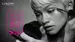 A person applying mascara while looking into a compact mirror, with text overlay promoting Lancome's Monsieur Big mascara reviews.