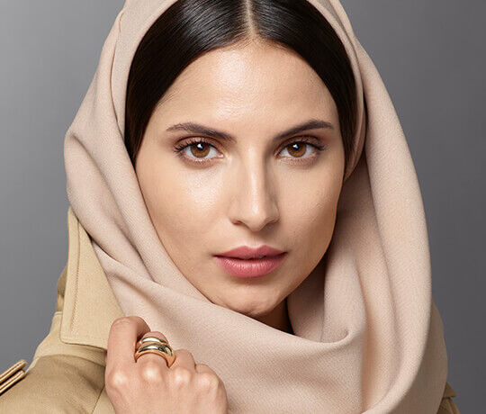 A close-up portrait of a woman wearing a light-colored hijab, with a neutral expression, showcasing her brown eyes and subtle makeup. She holds her hand near her face, displaying a few rings.