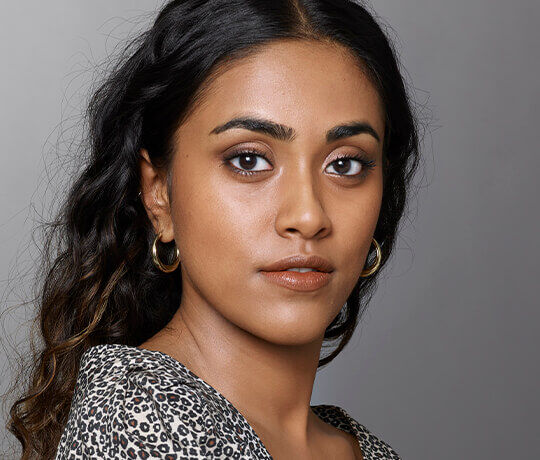 A close-up portrait of a young woman with long, dark hair, wearing hoop earrings and a patterned blouse. She has a natural makeup look, with a soft expression and a neutral background.