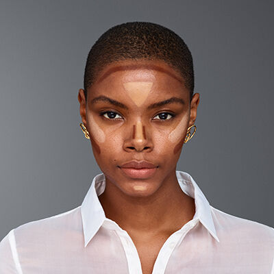 A close-up portrait of a woman with short, dark hair, showcasing a makeup look with contouring on her face. She is wearing a white button-up shirt and gold earrings, set against a neutral gray background.