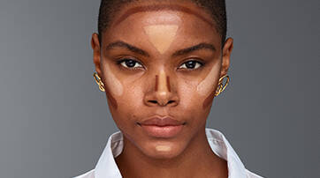 A close-up portrait of a woman with short, dark hair, showcasing a makeup look with contouring on her face. She is wearing a white button-up shirt and gold earrings, set against a neutral gray background.