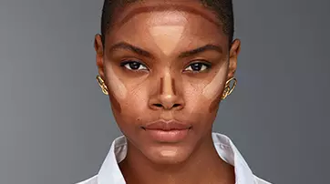 A close-up portrait of a woman with short, dark hair, showcasing a makeup look with contouring on her face. She is wearing a white button-up shirt and gold earrings, set against a neutral gray background.