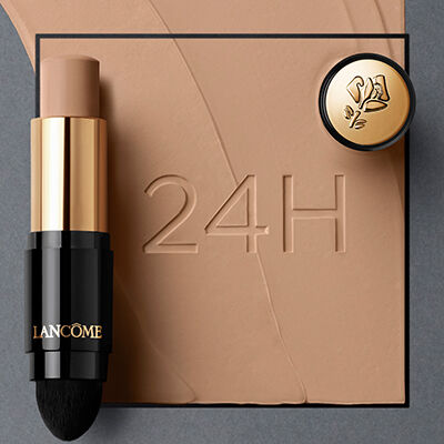 A stick foundation by Lancome is positioned beside a small round compact, set against a smooth, beige background with the text "24H" embossed in it.