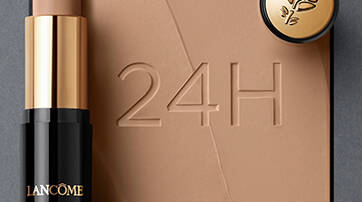 A stick foundation by Lancome is positioned beside a small round compact, set against a smooth, beige background with the text "24H" embossed in it.