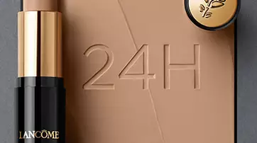 A stick foundation by Lancome is positioned beside a small round compact, set against a smooth, beige background with the text "24H" embossed in it.