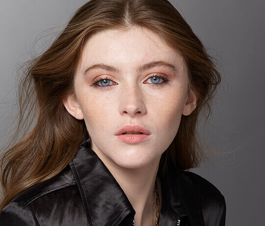 A close-up portrait of a young woman with long, wavy brown hair and blue eyes. She has a natural makeup look, featuring soft blush and muted lip color. She is wearing a black jacket and has a neutral expression. The background is light gray.