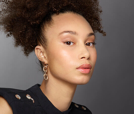 A close-up portrait of a young woman with curly hair, wearing a dark shirt and large gold earrings. She has a natural makeup look, with a soft pink lip color and a glowing complexion.