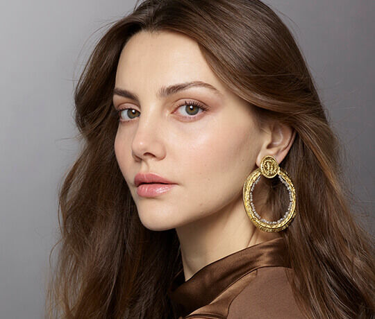 A woman with long, wavy brown hair and natural makeup wears large golden hoop earrings. She gazes directly at the camera with a subtle expression.