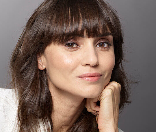 A close-up portrait of a woman with long, dark hair and a fringe, resting her chin on her hand and smiling gently. She has a natural, fresh makeup look and warm, expressive eyes, set against a soft gray background.