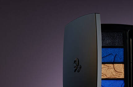 A stylish arrangement of a black eyeshadow palette featuring blue and gold shades, alongside a black eyeliner and a white mascara tube, set against a gradient background.