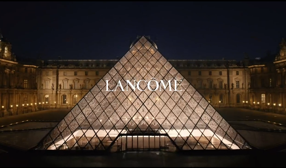 Une vue nocturne de la pyramide du Louvre illuminee, avec le mot "LANCOME" inscrit dessus.