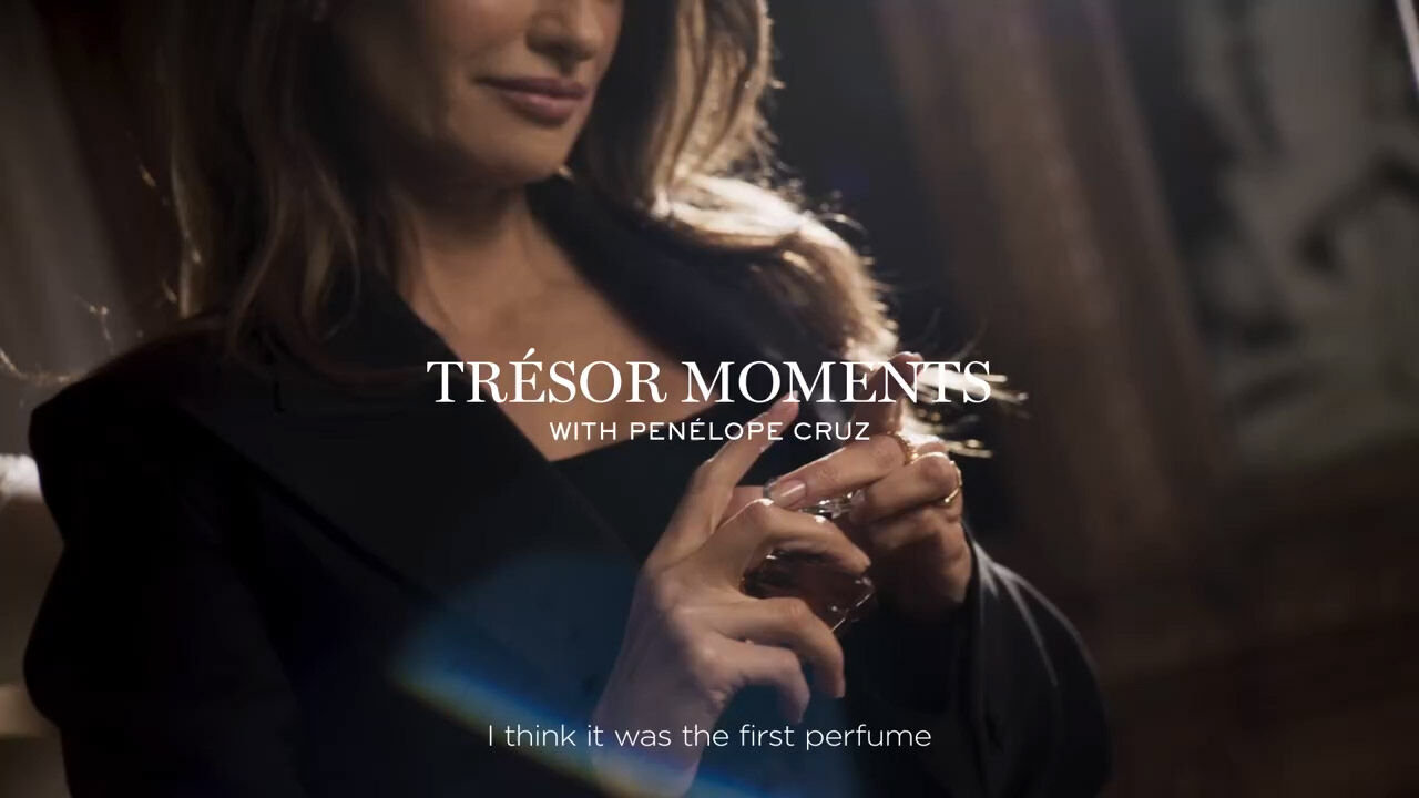A woman with long, wavy hair in a stylish black outfit holds a perfume bottle, with text overlay that reads "TRESOR MOMENTS WITH PENELOPE CRUZ." The caption below references a memory about the perfume.