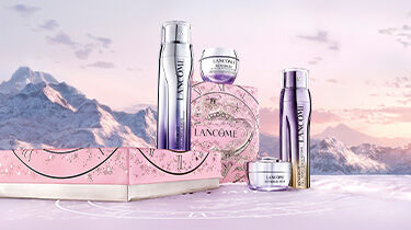 A stylish arrangement of three golden skincare products in a gift box, set against a dark, decorative background featuring intricate designs. Hands are gently holding the box.