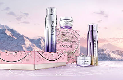 A stylish arrangement of three golden skincare products in a gift box, set against a dark, decorative background featuring intricate designs. Hands are gently holding the box.