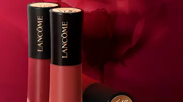 Two tubes of Lancome lipstick in varying shades of red are displayed in front of a blurred background featuring a rose. The tubes have sleek black caps with gold accents.