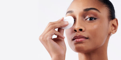 A woman is gently holding a cotton pad to her eye, demonstrating a skincare routine. She has a natural makeup look and a serene expression.