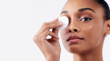 A woman is gently holding a cotton pad to her eye, demonstrating a skincare routine. She has a natural makeup look and a serene expression.