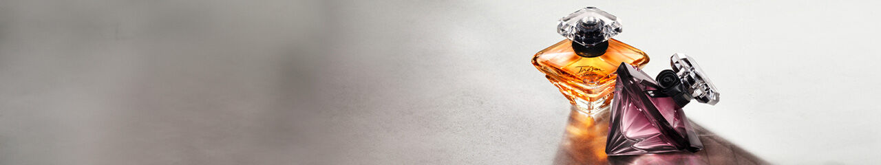 Two elegant perfume bottles are displayed on a smooth surface. The bottle on the left is amber-colored with a classic design, while the one on the right is a sleek, dark purple pyramid. Soft lighting highlights their shapes and colors.