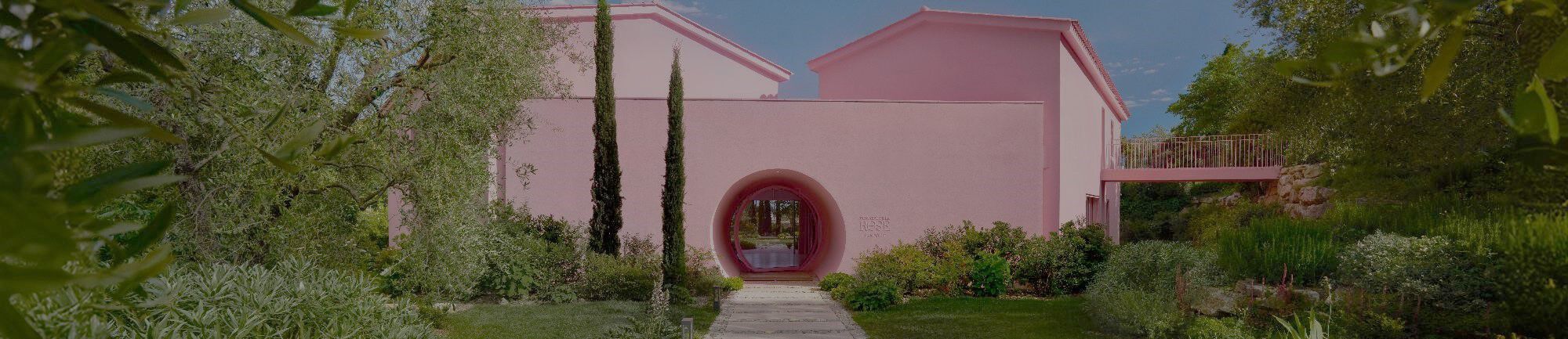 A pink stucco house surrounded by lush greenery, including trees and plants, with a curved entryway leading to a garden pathway.