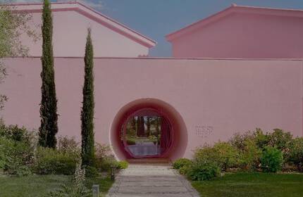 A pink stucco house surrounded by lush greenery, including trees and plants, with a curved entryway leading to a garden pathway.