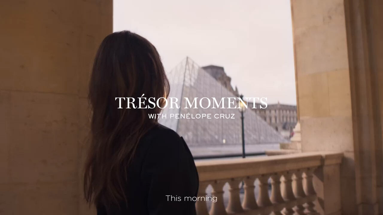 Une femme se tient pres de balustrade, regardant la pyramide du Louvre a Paris. Le texte indique "TRESOR MOMENTS WITH PENELOPE CRUZ" et "This morning".