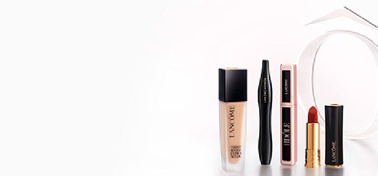 A display of three makeup products against a gradient brown background: a black eyeliner pencil, a beige foundation bottle, and a red lipstick tube, all branded with the Lancome logo.