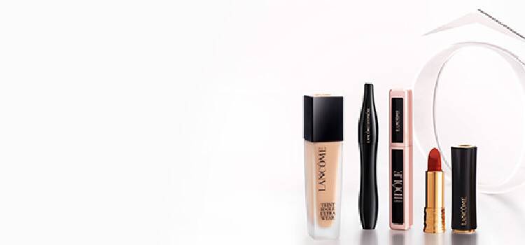 A display of three makeup products against a gradient brown background: a black eyeliner pencil, a beige foundation bottle, and a red lipstick tube, all branded with the Lancome logo.