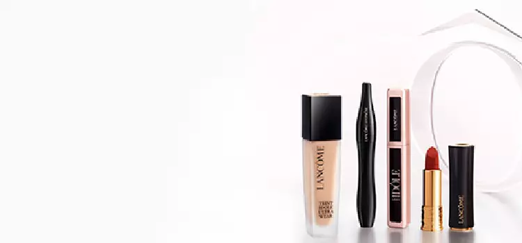 A display of three makeup products against a gradient brown background: a black eyeliner pencil, a beige foundation bottle, and a red lipstick tube, all branded with the Lancome logo.