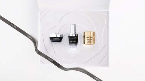 Boite blanche ouverte contenant trois produits de soin Lancome, sur un fond blanc, accompagnee d'un ruban portant la marque.