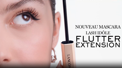 A close-up of an eye showcasing long, defined eyelashes. A mascara wand is positioned next to the eye, with promotional text for Lancome's "Lash Idole Flutter Extension" mascara.
