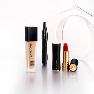 A collection of Lancome cosmetics, including a foundation bottle, a mascara, and two lipsticks—one red and one in a black tube—arranged on a reflective surface with a white circular backdrop.