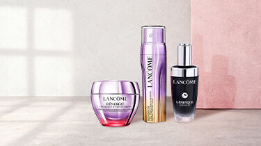 Three Lancome skincare products are displayed against a soft gradient background. The products include a black serum, a blue jar labeled "Renergie H.P.N. 300-Peptide Cream," and a gold jar labeled "Absolue Soft Cream."