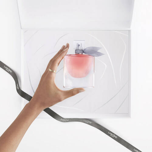 Une main tient un flacon de parfum avec un degrade rose, pose sur un fond blanc. Un ruban gris avec le logo "Lancome" est visible sur le cote.