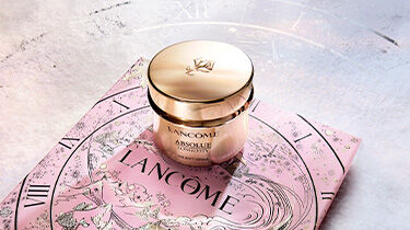 Three luxurious skincare products from Lancome are showcased against a dark background, featuring elegant gold packaging. The items include a serum and two cream jars, highlighting sophistication and beauty.