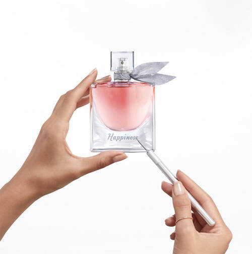 Une main tient un flacon de parfum en verre transparent avec un ruban gris, marque "Happiness", tandis qu'une autre main utilise un outil pour le decorer.