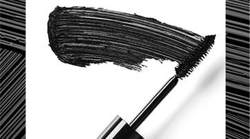 A close-up of a mascara wand coated with black mascara, with a smudge of mascara alongside it on a white background.