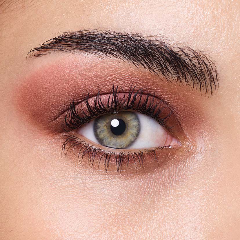 A close-up of an eye featuring a light hazel iris with a dark pupil, complemented by soft, blended eyeshadow in warm tones and well-defined eyelashes.
