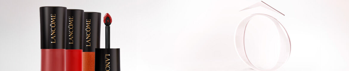 Three Lancome lipstick tubes in various shades, with one applicator prominently displayed, set against a light background featuring a circular design.