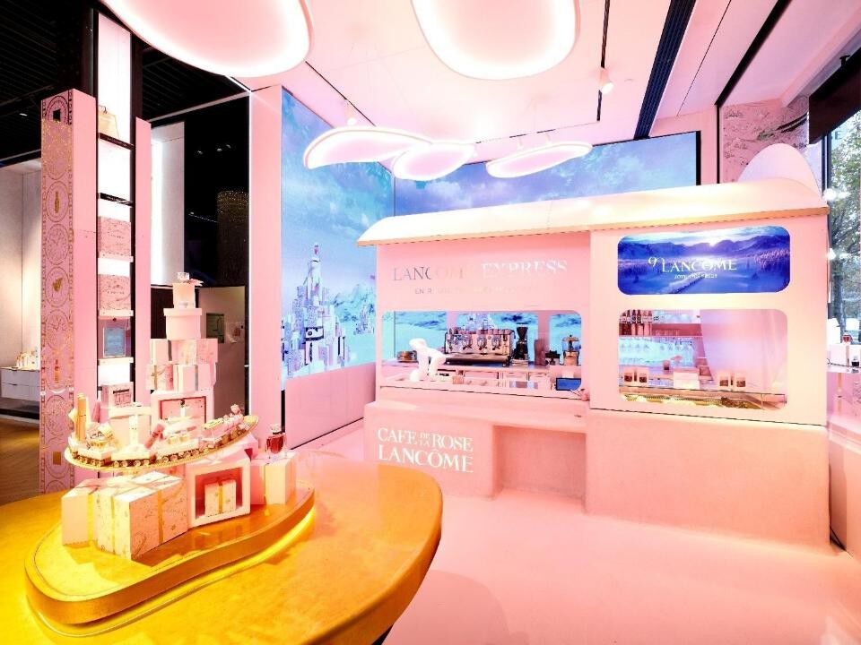 Vue interieure d'un magasin Lancome avec un comptoir sur le theme d'un train rose, une presentation de produits disposes en tour de cadeaux et de grands ecrans numeriques affichant des arriere-plans sceniques.