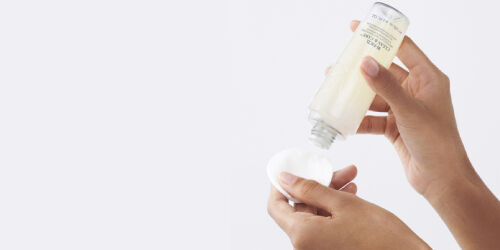 A person’s hand is holding a cotton pad while pouring a skincare product from a bottle onto it.