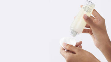 A person’s hand is holding a cotton pad while pouring a skincare product from a bottle onto it.