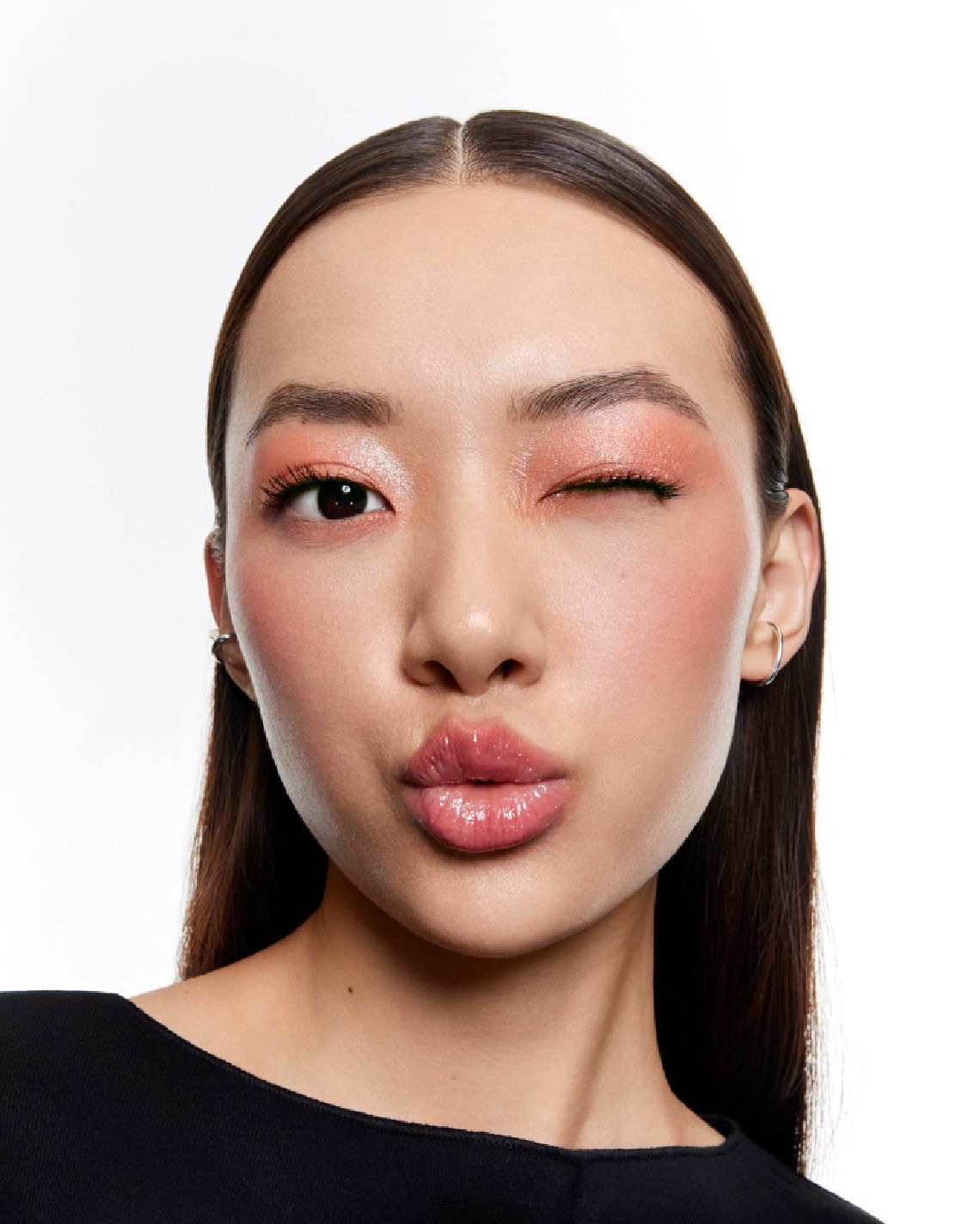 Femme faisant un clin d'oeil et un bisou, avec un maquillage corail scintillant et des levres roses brillantes.