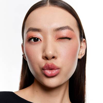 Femme faisant un clin d'oeil et un bisou, avec un maquillage corail scintillant et des levres roses brillantes.