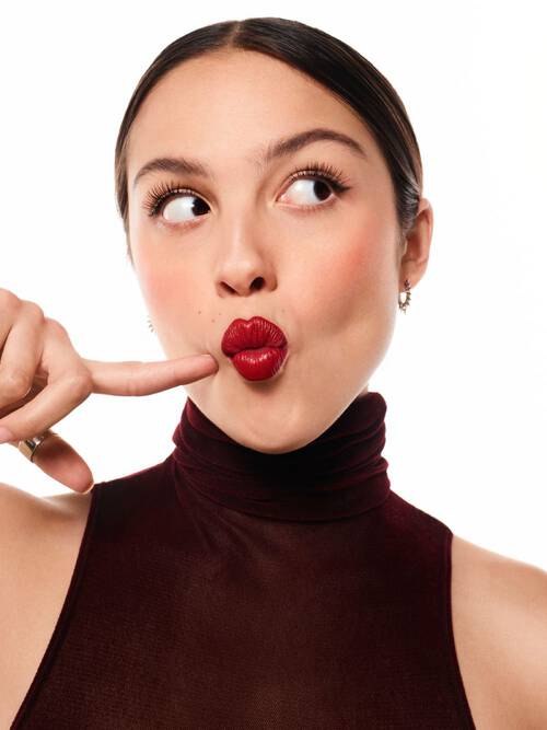 A woman in a burgundy turtleneck playfully makes a quirky expression, pressing her finger to her lips, with shiny red lipstick on and a light background.