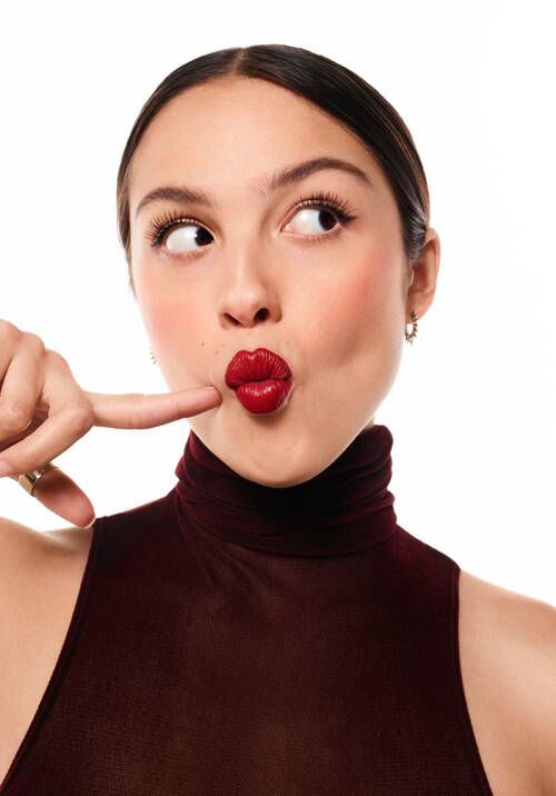 A woman with brown hair styled in a sleek bun is playfully holding her finger to her lips, making a kissy face with vibrant red lipstick, wearing a burgundy high-neck top, against a white background.