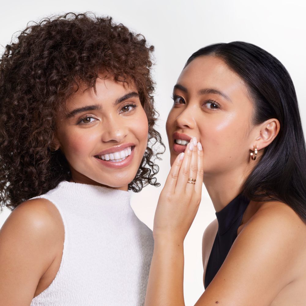 Deux femmes souriantes posent ensemble. L'une a des cheveux boucles et porte un haut blanc, tandis que l'autre a des cheveux lisses et un haut noir, chuchotant avec un sourire complice.